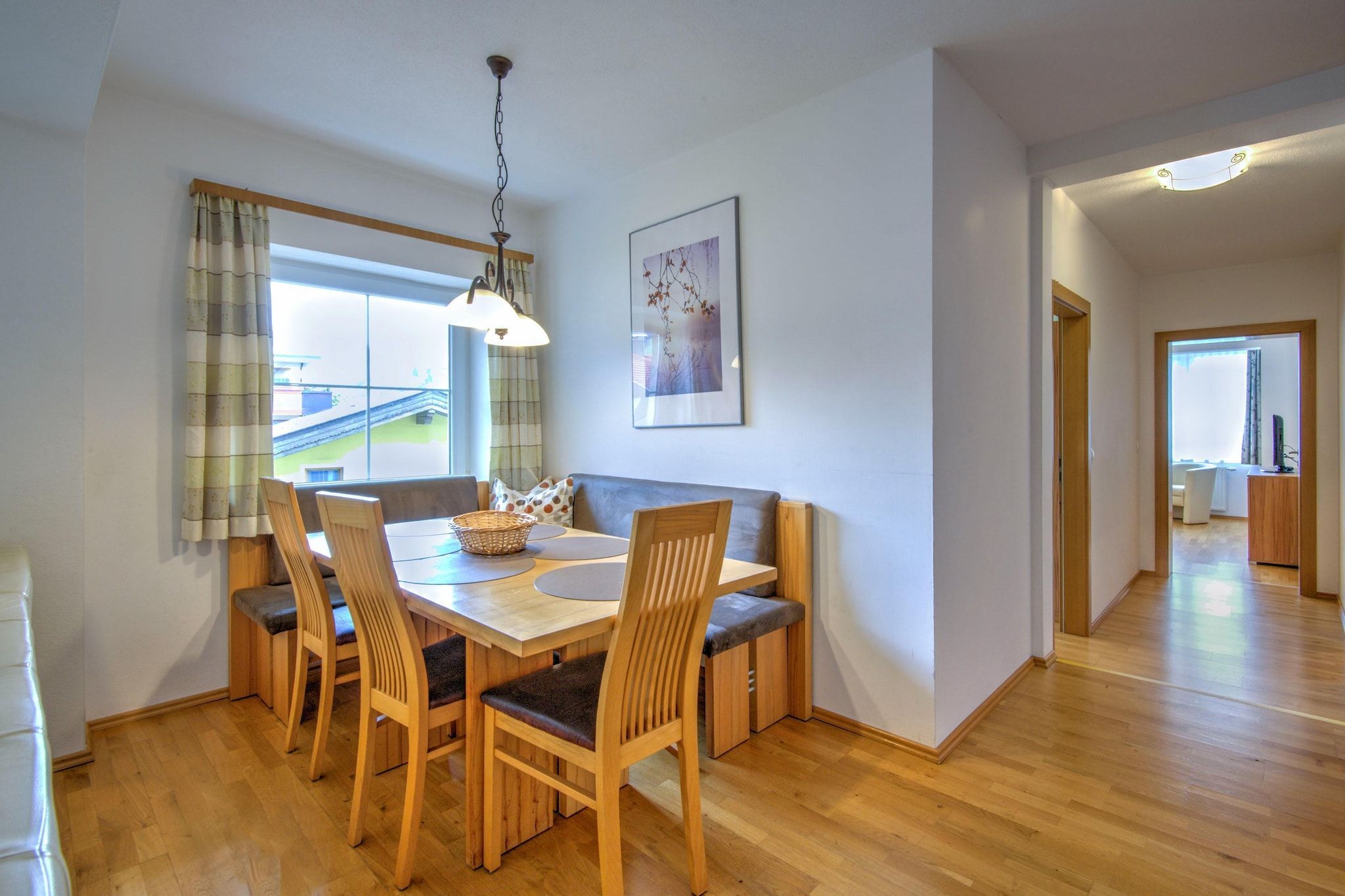 A dining area with a wooden table, chairs, and a bench. There is a window with curtains, a hanging light fixture, and a framed picture on the wall.
