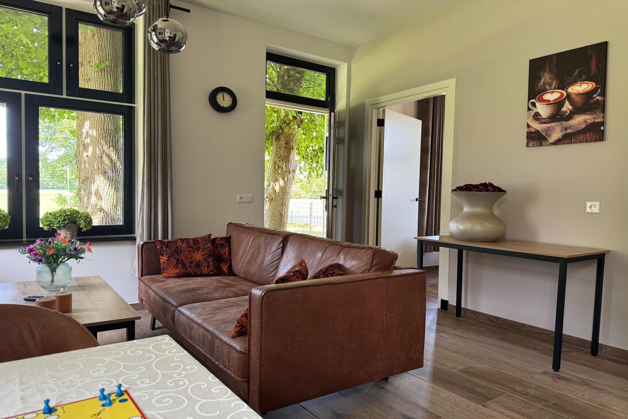 A living room with a brown leather sofa, a wooden coffee table, a vase with flowers, and a large window with curtains.