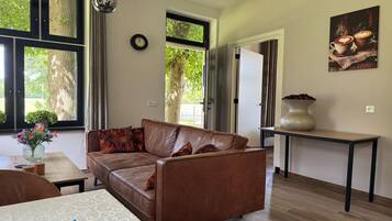A living room with a brown leather sofa, a wooden coffee table, a vase with flowers, and a large window with curtains.
