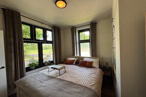 A bedroom with a bed, a bedside table, a window with curtains, and a view of greenery outside. - Langelshoes Guesthouse (Haaksbergen)
