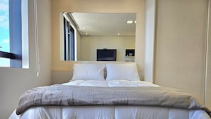 Cama bem arrumada com edredom acolchoado e travesseiros, mesa de cabeceira com abajur e janela com cortinas.