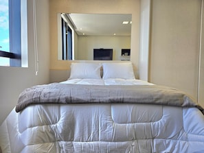 Una cama bien hecha con edredón acolchado y almohadas, una mesita de noche con lámpara, y una ventana con cortinas.