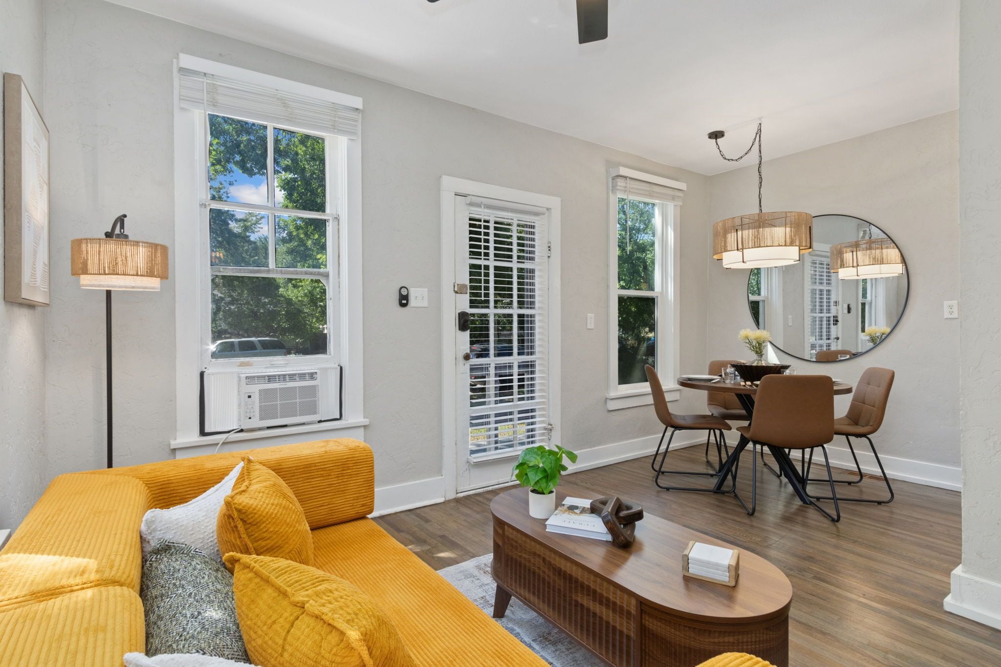 A modern living room with a yellow sofa, a dining area with a table and chairs, and large windows with blinds.