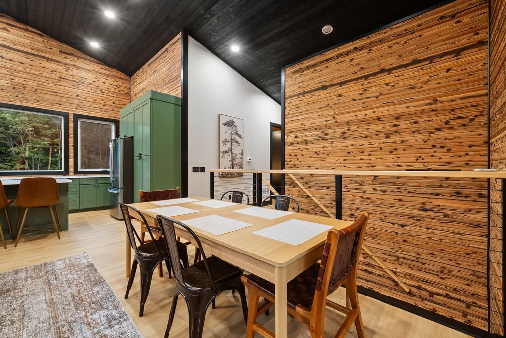 A modern interior with a dining area featuring a wooden table and chairs, a kitchen with green cabinets, and a view of the outdoors through large windows.