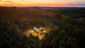 A modern house surrounded by dense forest, with a well-lit interior visible from the outside. - Escape to Nature's Wonderland: The rolling  foothills of Appalachia (Logan)