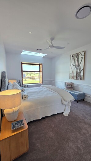 Een slaapkamer met een bed, nachtkastje, lamp, plafondventilator, raam en een schilderij.