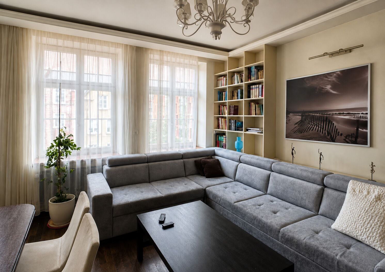 Un salon moderne avec un canapé d’angle, une table basse, une bibliothèque et une grande fenêtre agrémentée de rideaux légers.