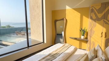 Une chambre d’hôtel avec une grande fenêtre, un lit avec du linge de maison blanc, un miroir et une petite plante posée sur une étagère.