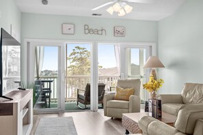A living room with a sofa, armchair, and a table with a lamp. There is a sliding glass door leading to a balcony with outdoor furniture. - 10% off 3-7 nights thru Dec. 2BR 2BA condo. Lake view, gated comm. Short walk to beach. (North Myrtle Beach)