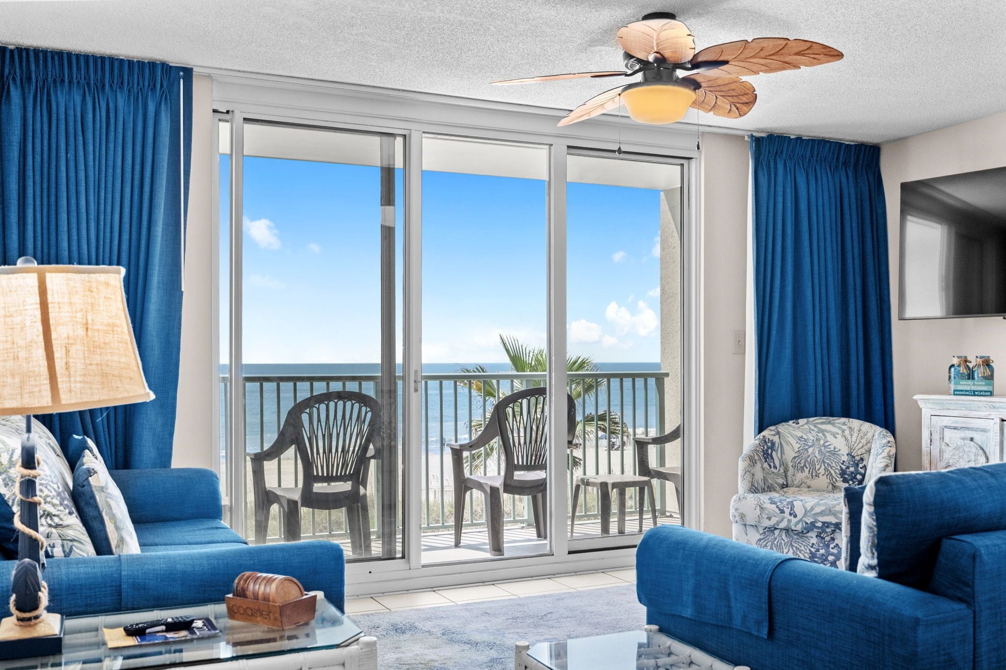 Une pièce dotée d’une grande porte-fenêtre coulissante donnant sur un balcon avec vue sur l’océan. On y trouve deux canapés bleus, une table basse et un ventilateur de plafond.