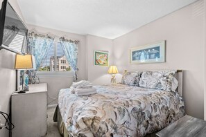 Ein Schlafzimmer mit einem Bett, zwei Nachttischlampen, einem Fenster mit Vorhängen, einem Wandbild und einem kleinen Tisch mit Lampe.