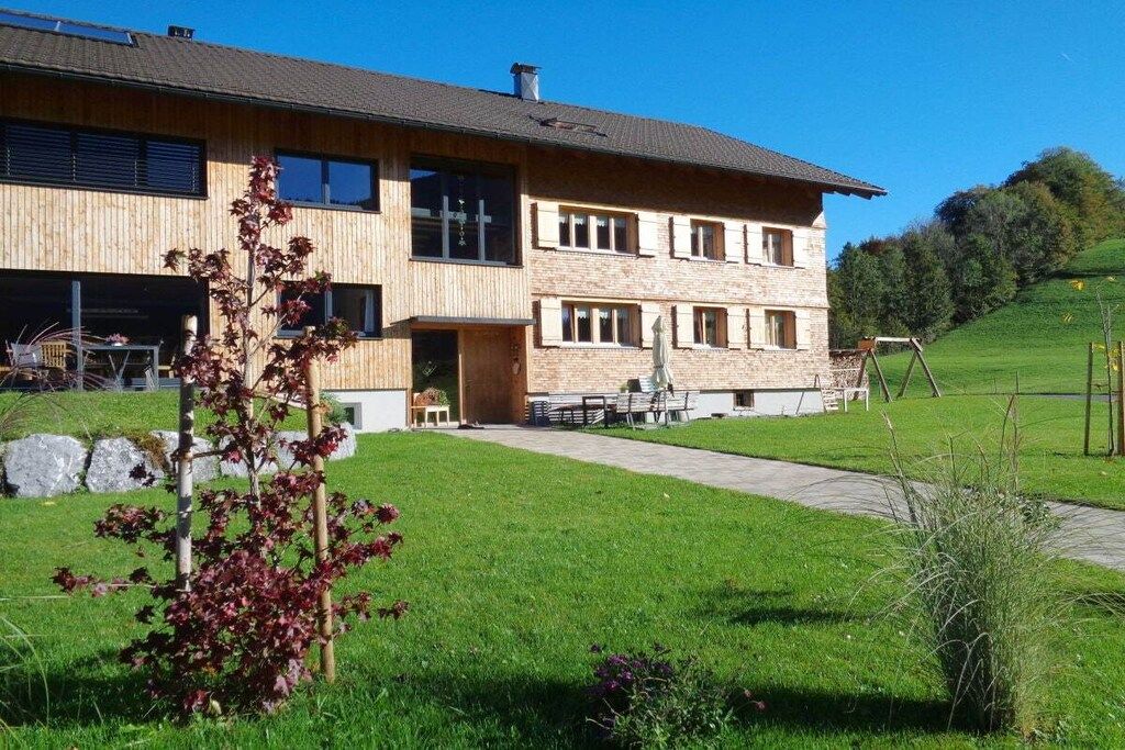 Un bâtiment à deux étages, doté d’une façade en bois et de quelques éléments en pierre, une allée pavée et une pelouse verdoyante avec un petit arbre.