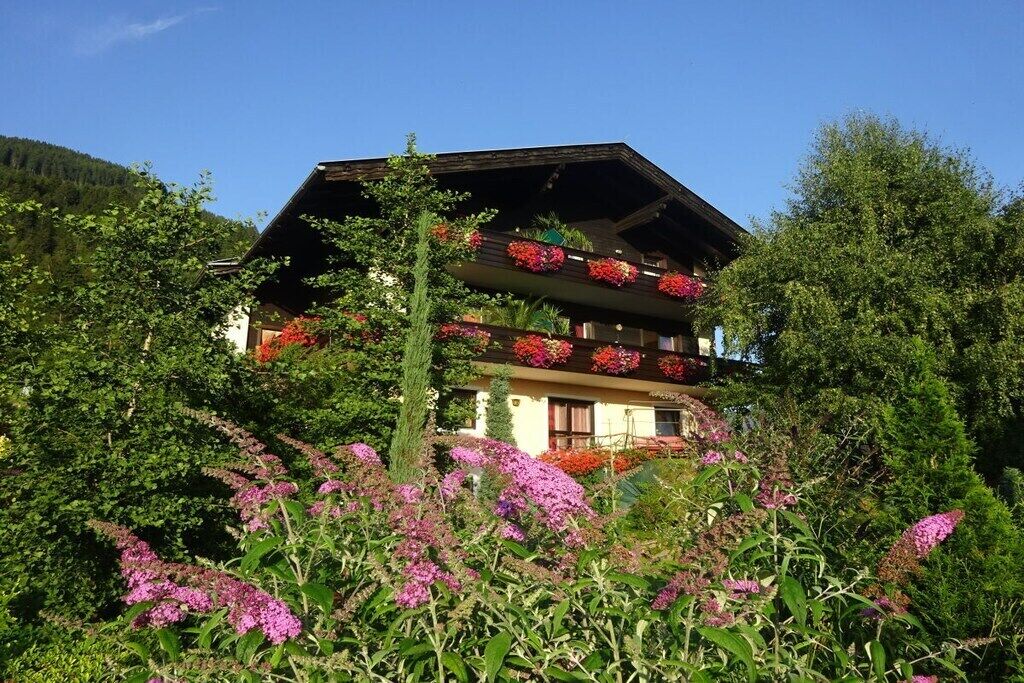 Eine zweigeschossige Chalet mit Balkonen, umgeben von üppigem Grün und blühenden Pflanzen.