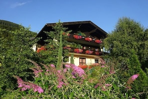 Eine zweigeschossige Chalet mit Balkonen, umgeben von üppigem Grün und blühenden Pflanzen.