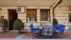 Un espacio al aire libre con mesas y sillas, una planta en maceta y la entrada de un edificio con puerta y ventanas.