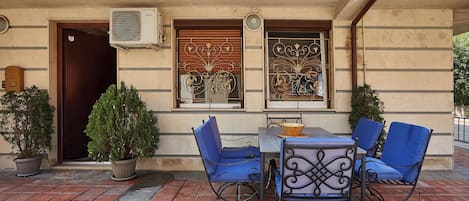 An outdoor seating area with a table and chairs, a potted plant, and a building entrance with a door and windows.