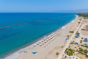 An aerial view of a beach with a long stretch of sand, a clear blue sea, and a few people. There are rows of beach umbrellas and sun loungers. - Aurelia Beach Residence by Nomads (Paphos)
