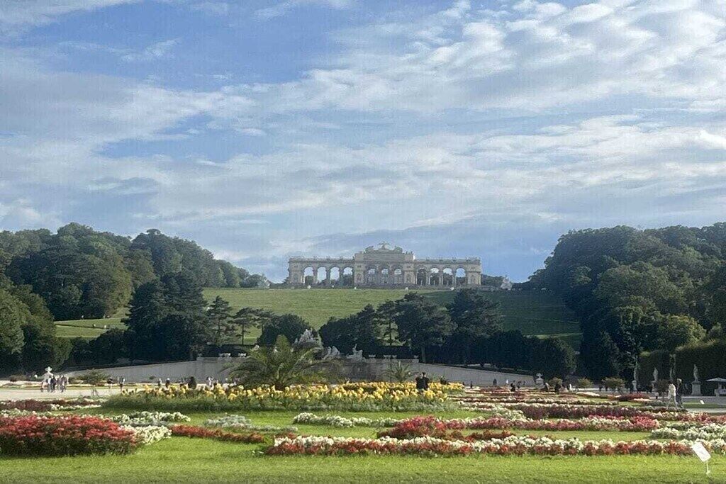 Un vaste parc ouvert, avec un bâtiment imposant en arrière-plan, entouré d’une végétation luxuriante et de parterres de fleurs.
