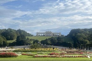 Um parque amplo e aberto, com um edifício imponente ao fundo, rodeado por vegetação exuberante e canteiros de flores.