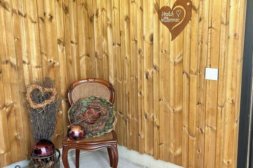 A wooden wall with a heart-shaped sign, a wicker chair with a decorative cushion, and a metal sculpture.