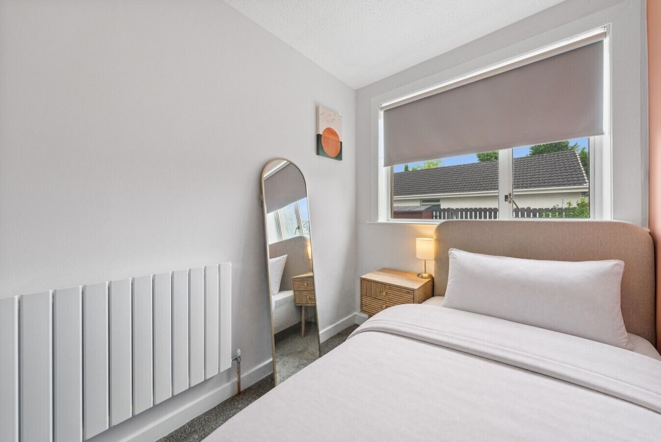 A hotel room with a bed, a bedside table with a lamp, a window with a view of a house, a radiator, and a mirror.