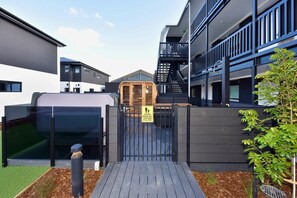 A gated entrance to a modern building with a wooden door, a sign indicating a child-friendly area, and a small garden area with mulch and a tree. - The Wellness Nook for a Relaxing City Stay (Christchurch)