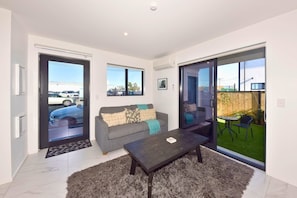 A modern living room with a sofa, a coffee table, and a sliding glass door leading to a patio with outdoor furniture. - The Modern Hideaway for Every Stay (Christchurch)