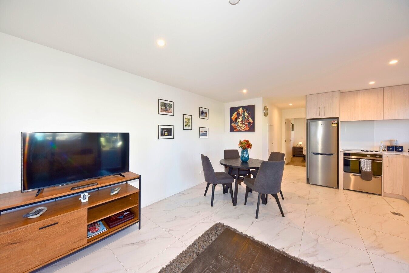 A modern living room with a flat-screen TV, wooden entertainment unit, dining table with chairs, and a kitchen area with a refrigerator and oven.