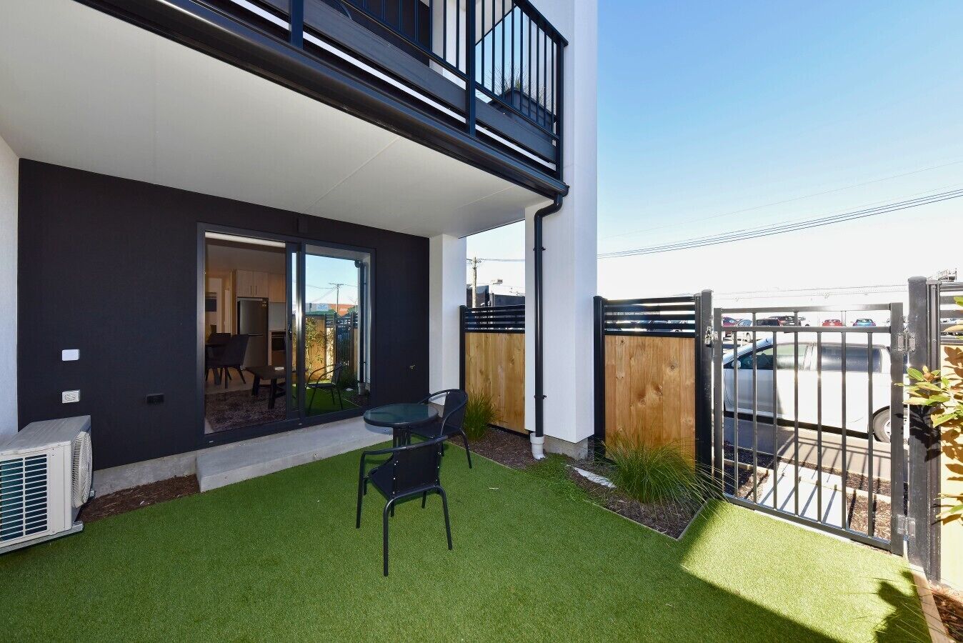 A modern building with a balcony, a small patio with artificial grass, and a view of a parking area.