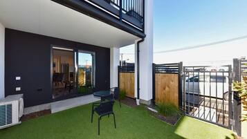 A modern building with a balcony, a small patio with artificial grass, and a view of a parking area.
