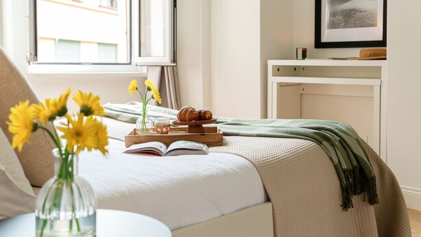 Quarto bem arrumado com cama, mesa de cabeceira com um livro, vaso com flores e bandeja com doces.