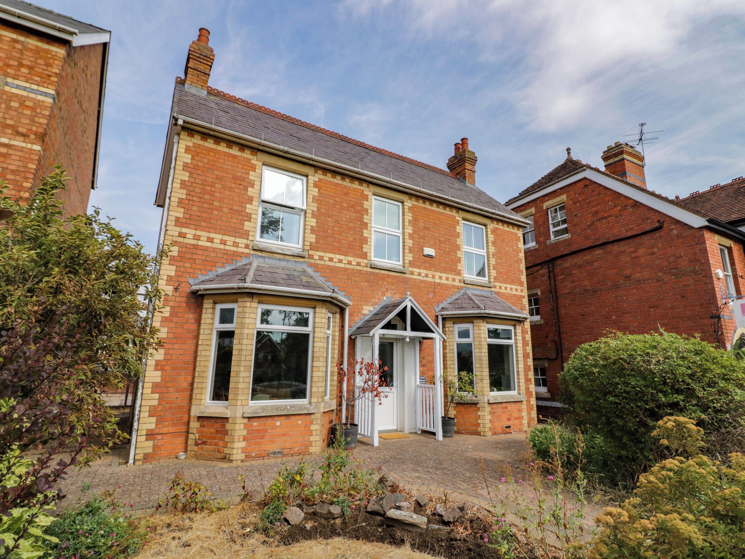 A two-story brick house with a front porch, a bay window, and a small garden area.