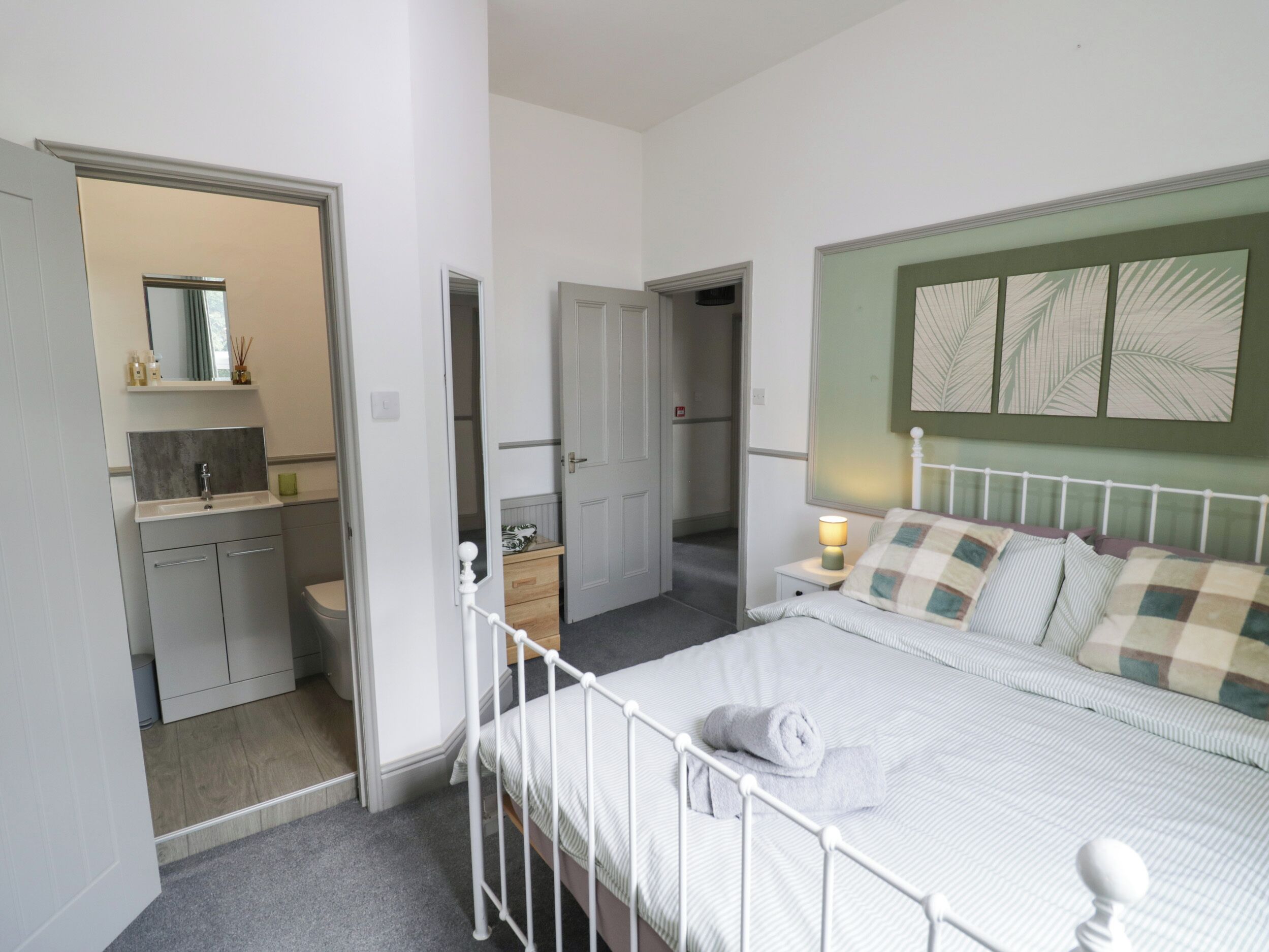 A bedroom with a bed, bedside table, and a nightstand. There is a bathroom with a sink and mirror. The room has a door leading to another room.