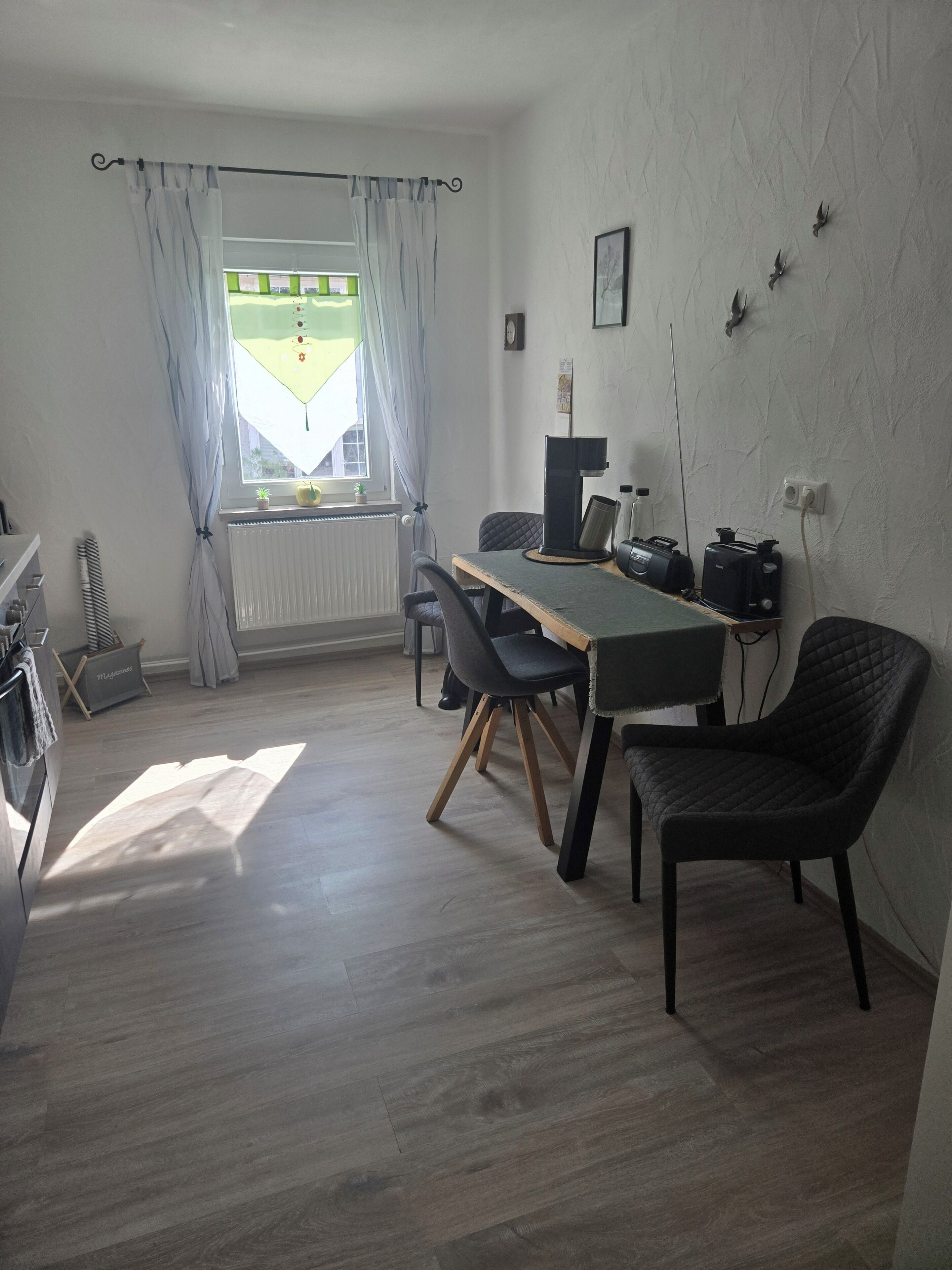 Una habitación con una mesa de comedor, sillas, un radiador, una ventana con cortinas, un reloj de pared y una estantería de pared con objetos.