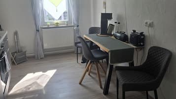 Una habitación con una mesa de comedor, sillas, un radiador, una ventana con cortinas, un reloj de pared y una estantería de pared con objetos.