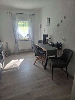 Una habitación con una mesa de comedor, sillas, un radiador, una ventana con cortinas, un reloj de pared y una estantería de pared con objetos.