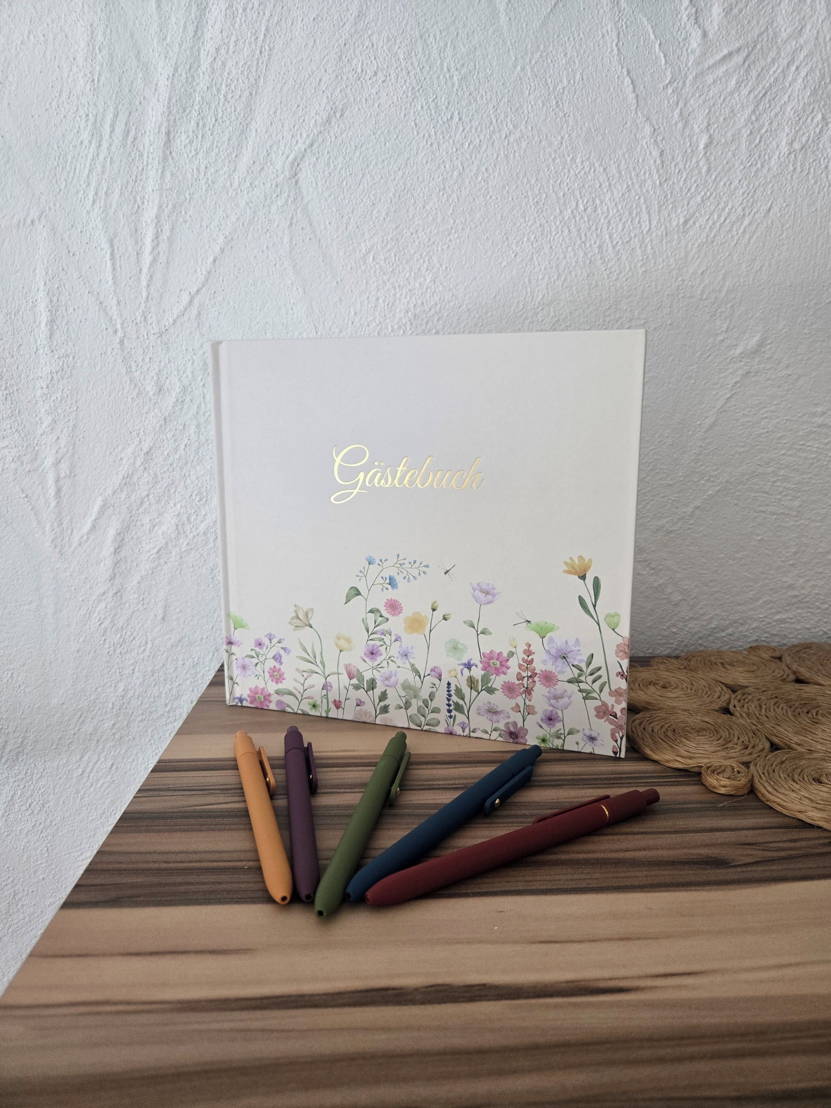 Tarjeta blanca con la palabra 'Gästebuch' escrita en dorado, sobre una superficie de madera con bolígrafos de colores a un lado.