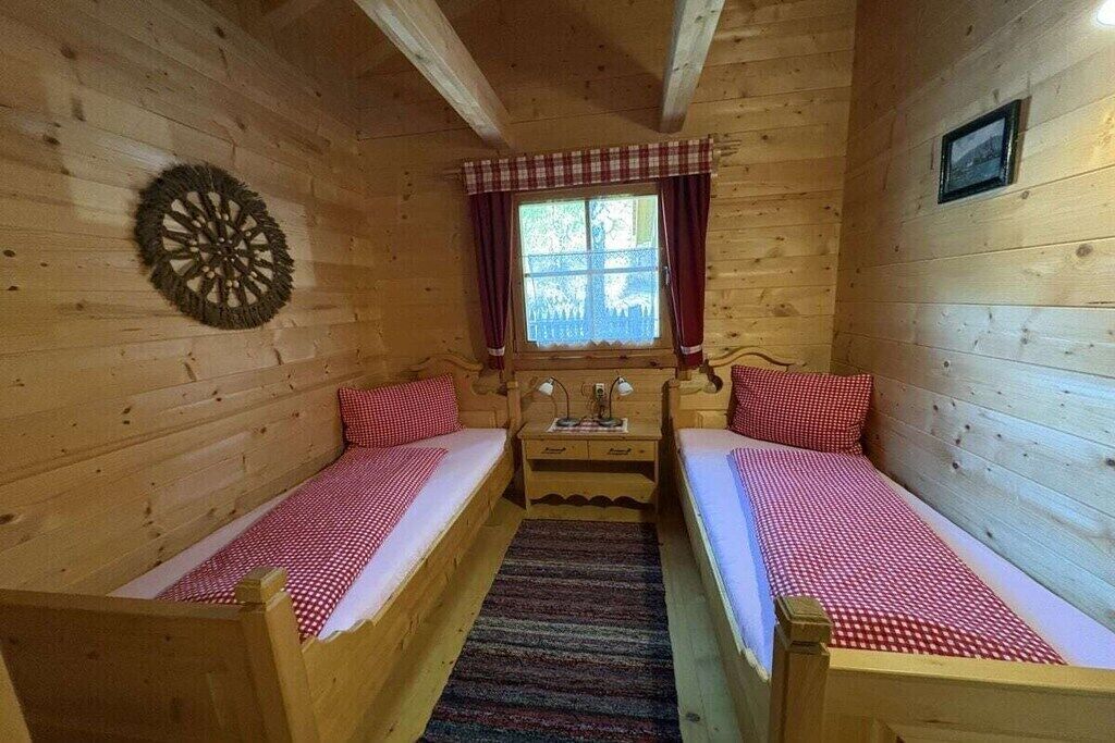 Une chambre dans un chalet en bois, avec deux lits simples, une petite table, une fenêtre avec des rideaux et un élément décoratif mural.