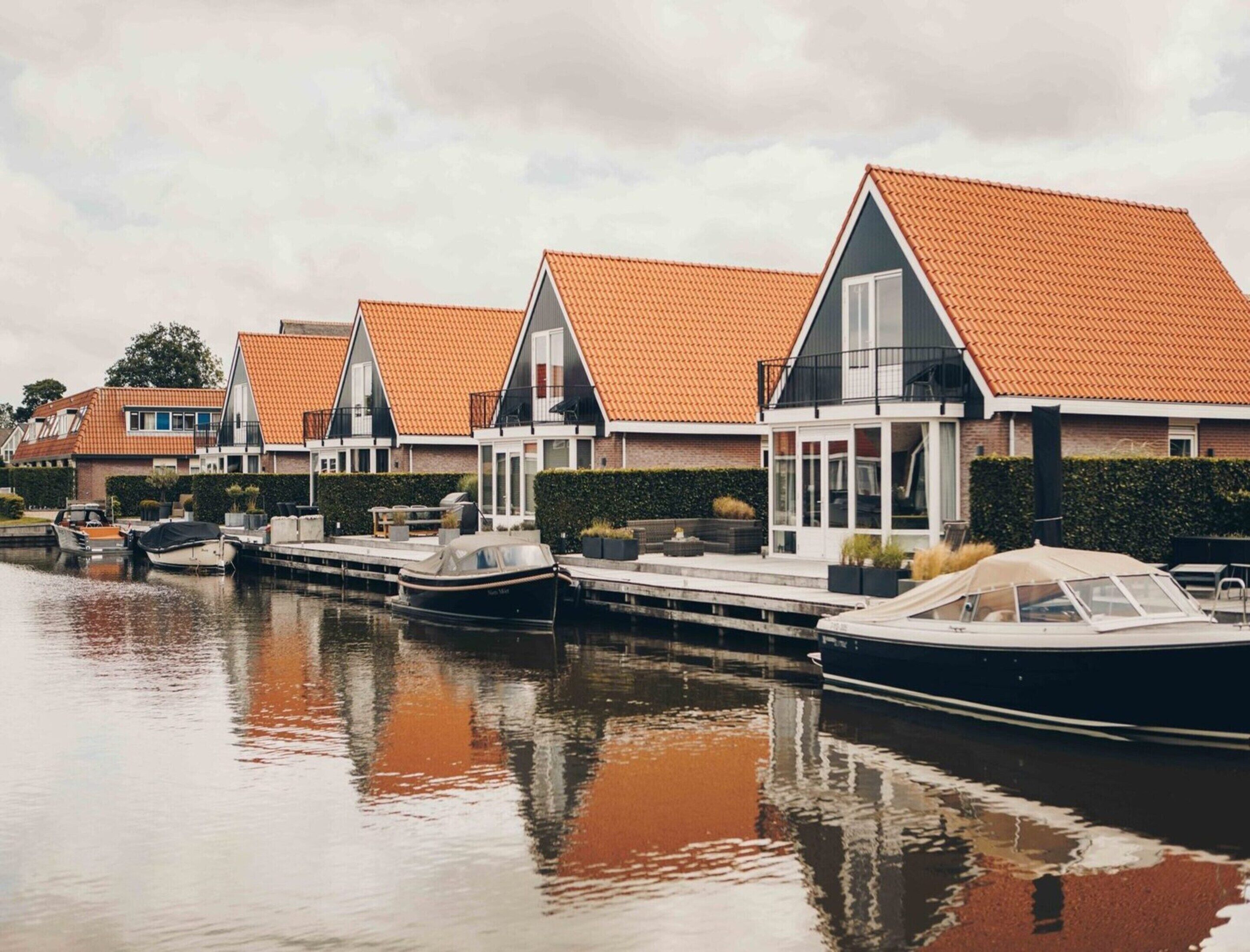 A row of modern houses with orange tiled roofs, a canal, and moored boats.