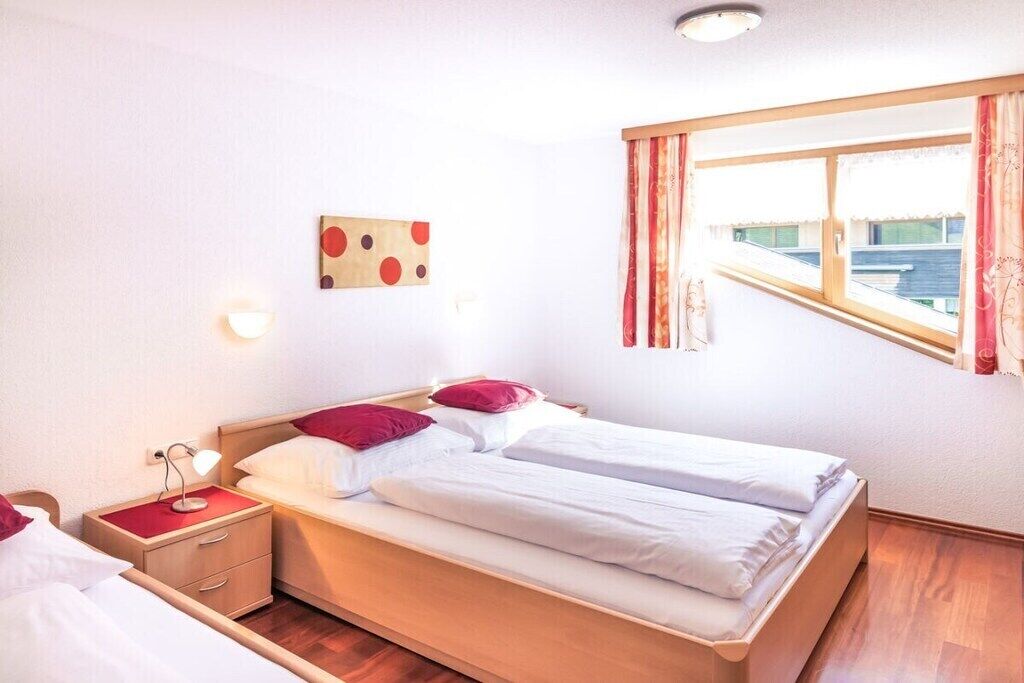 Een tweepersoonsbed met twee kussens, een nachtkastje met lamp, een wandklok en een raam met gordijnen.
