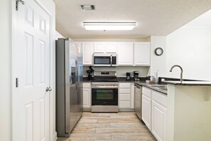 A modern kitchen with white cabinetry, stainless steel appliances, a granite countertop, and a wooden floor. - New Gulf-Front Listing| Amazing Views| Pools| Tennis Courts| Gym| Free Rounds of Golf (Pensacola)