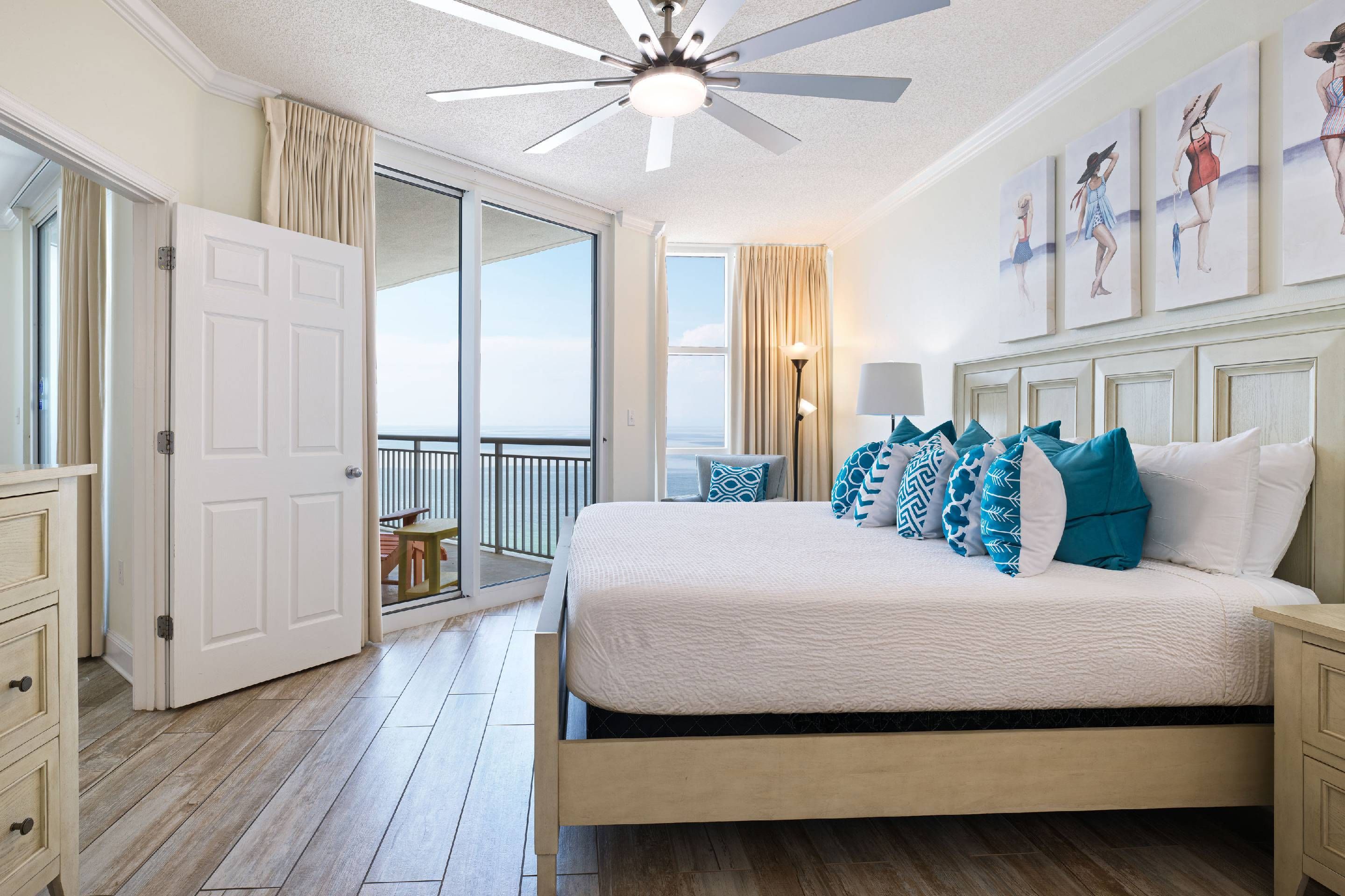 A bedroom with a bed, a ceiling fan, a view to the outside, and artwork on the wall.