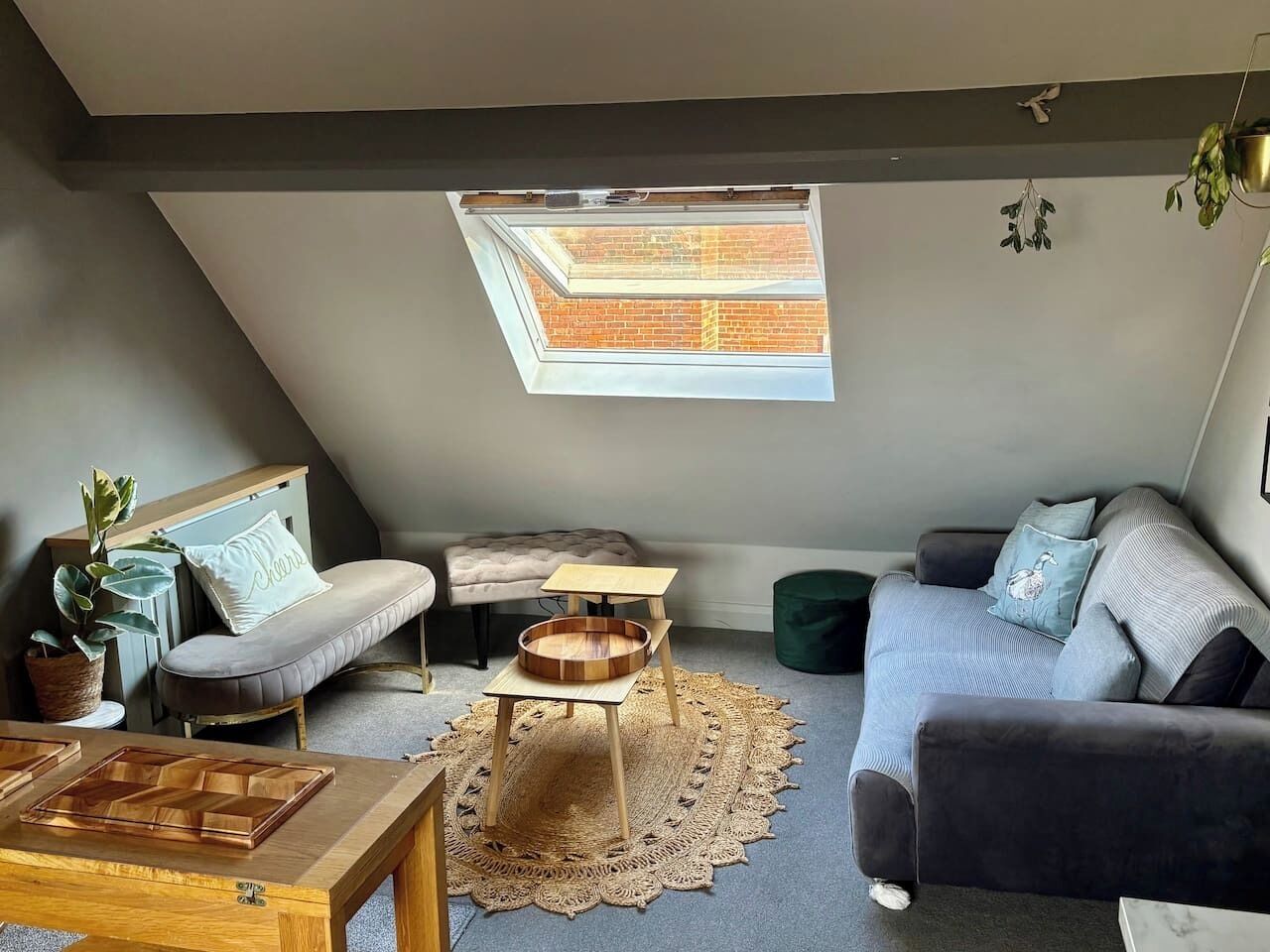 A cozy loft-style room with a sloped ceiling, a skylight, a grey sofa, a small wooden table, and a cushioned bench.