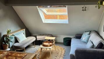 A cozy loft-style room with a sloped ceiling, a skylight, a grey sofa, a small wooden table, and a cushioned bench.