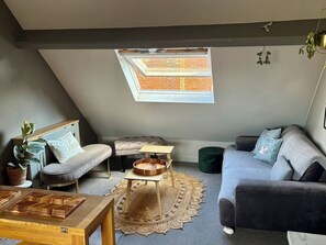 A cozy loft-style room with a sloped ceiling, a skylight, a grey sofa, a small wooden table, and a cushioned bench.