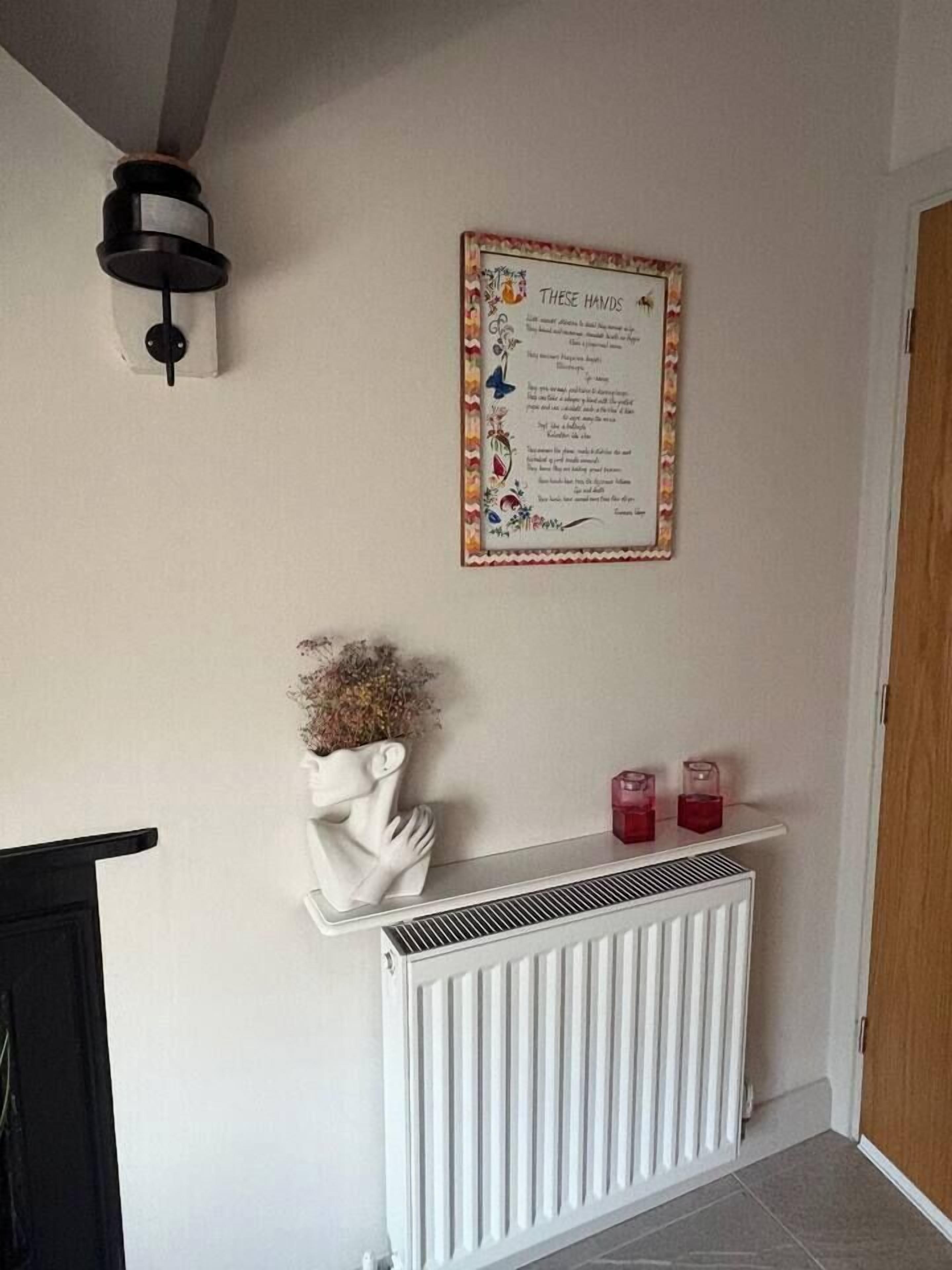Una habitación con un radiador blanco, una estantería blanca montada en la pared con adornos, y un texto enmarcado en la pared.