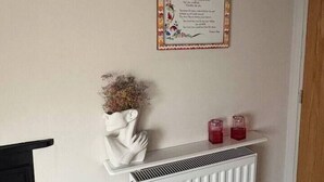 Une pièce avec un radiateur blanc, une étagère blanche fixée au mur, ornée d’objets décoratifs, et un texte encadré accroché au mur.