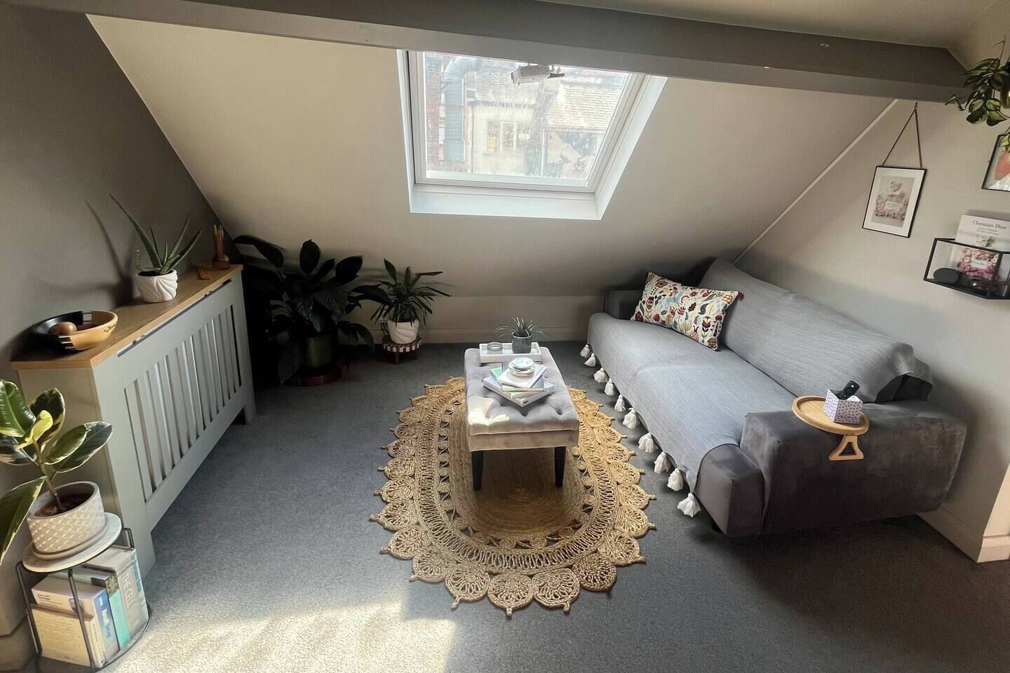 Ein gemütliches Schlafzimmer im Loft-Stil mit schrägem Dach, einem grauen Sofa, einem Couchtisch mit Büchern und mehreren Blumentöpfen.