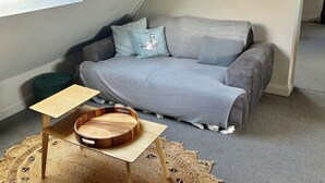 Une chambre à coucher aménagée en mezzanine, chaleureuse, avec un canapé gris, une petite table d’appoint en bois et un tapis tissé.