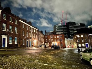 A nighttime urban street with brick buildings, a wet cobblestone road, and a modern building with cranes in the background. - Gracious Apartment in Historic Sheffield Georgian Quarter (Sheffield City Centre)
